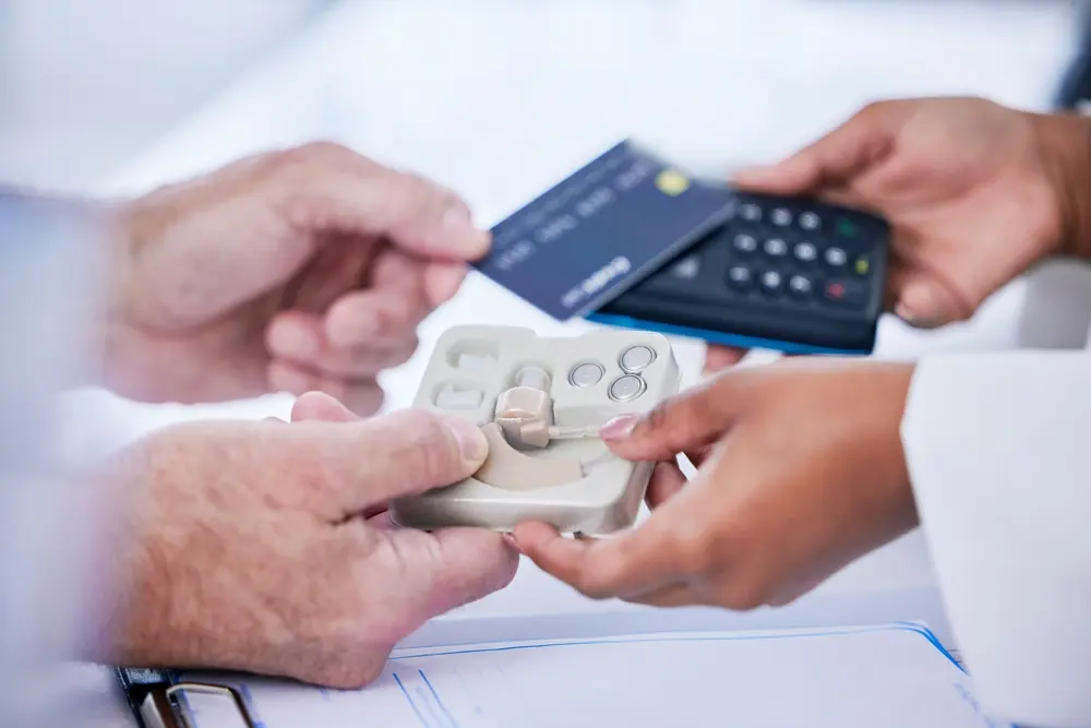 Two people exchange a payment card and card reader over a tray holding hearing aids and batteries, suggesting a purchase or payment for hearing aid devices.