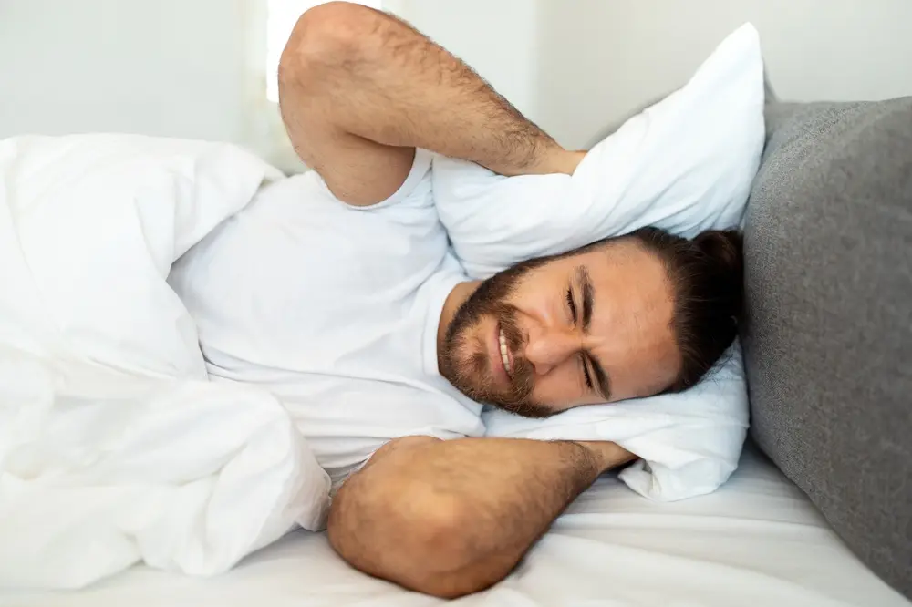 A man lying in bed covers his ears with a pillow, eyes closed and face tense, appearing disturbed or annoyed by noise while trying to sleep.