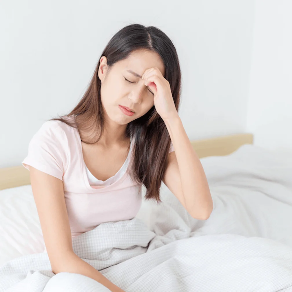 A woman sits on a bed with her eyes closed and one hand on her forehead, appearing to have a headache or feel unwell.