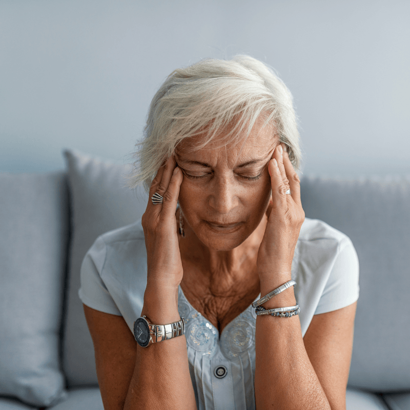 Older woman with short gray hair sits on a couch, eyes closed, touching her temples with both hands, appearing to have a headache or be in discomfort.
