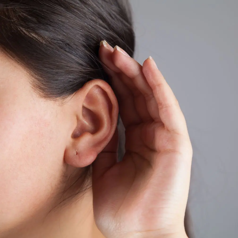 A person holds their hand behind their ear, as if trying to hear better. Only the ear, hand, and part of the face are visible, with a neutral gray background.