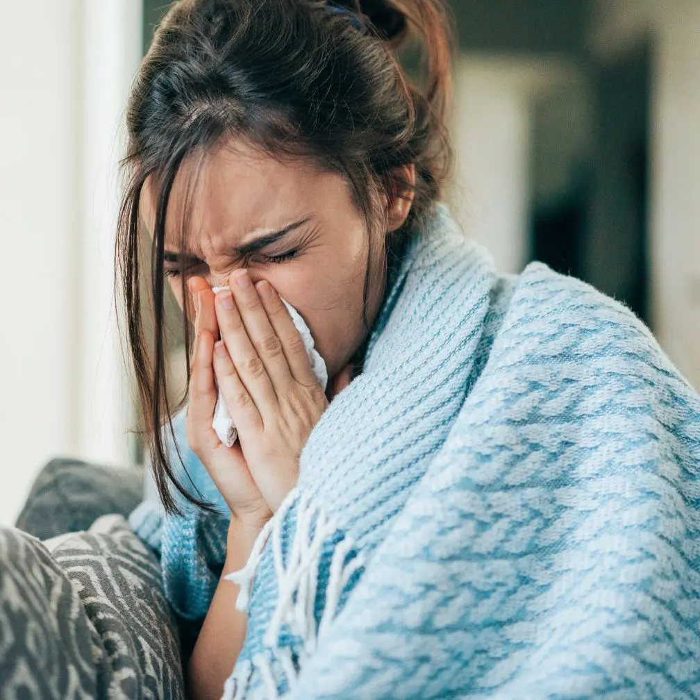 A woman wrapped in a blue blanket sits on a couch, holding a tissue to her nose as if sneezing or blowing her nose, appearing unwell.