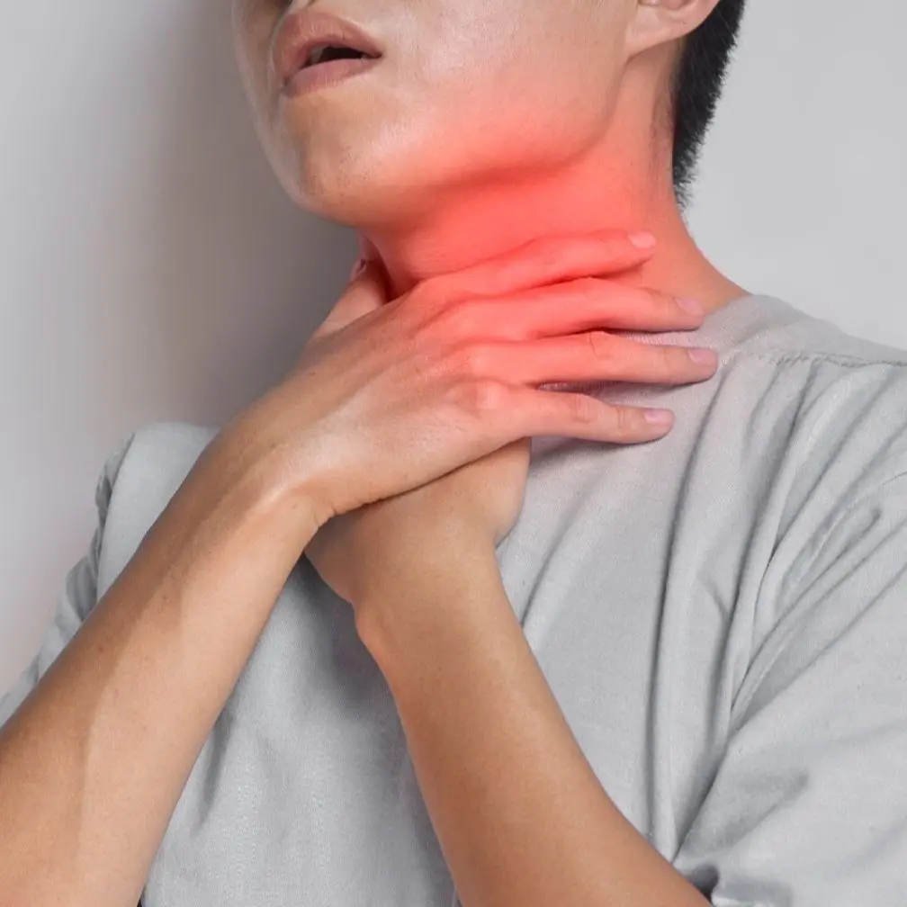 A person in a gray shirt touches their throat with both hands, showing discomfort. The neck area is highlighted in red, indicating pain or a sore throat.