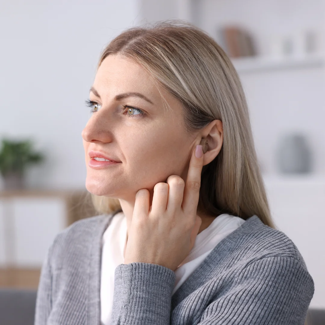 A woman with blonde hair touches her ear with her finger, appearing thoughtful. She is wearing a gray sweater and sitting indoors with blurred shelves and a plant in the background.