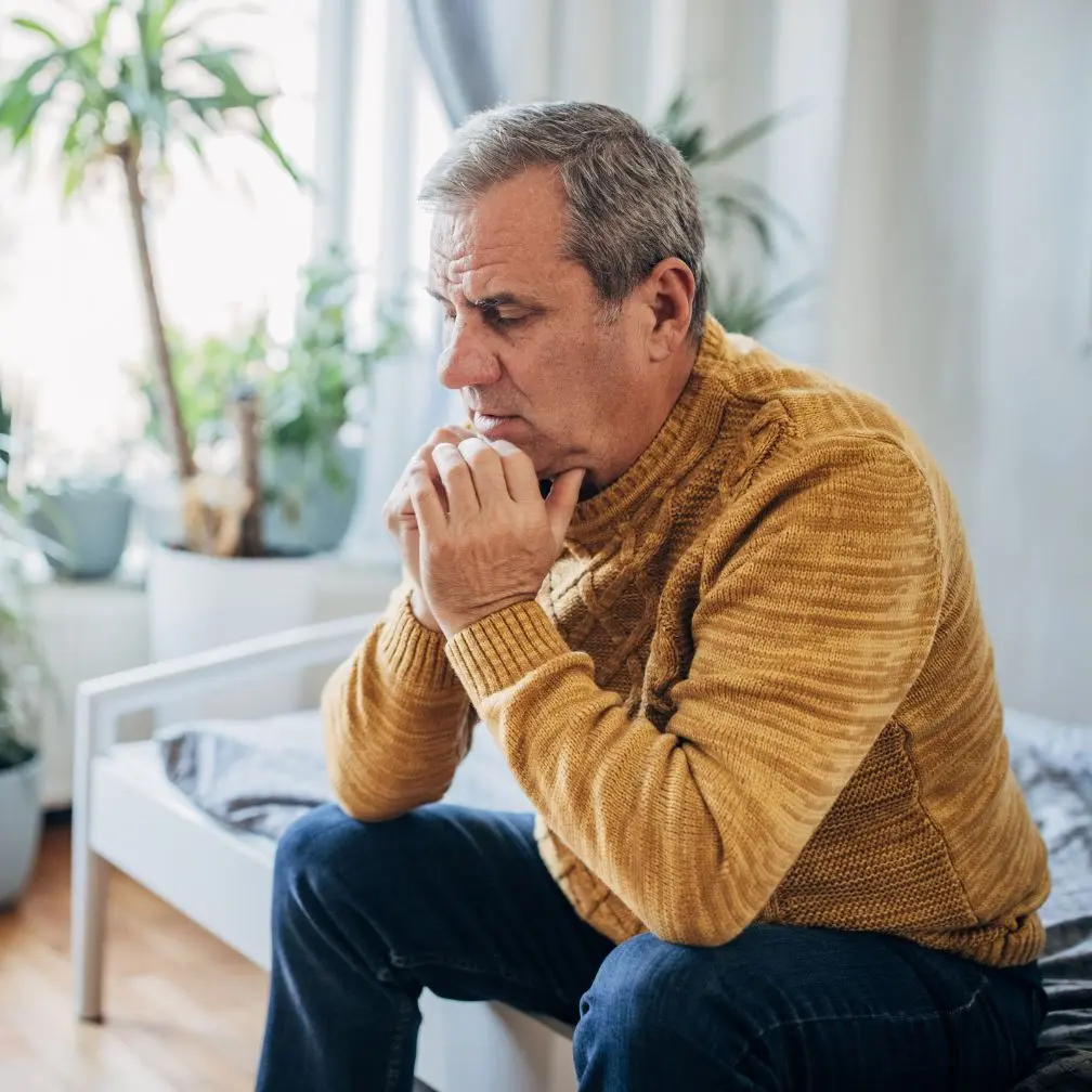 An older man in a yellow sweater sits on a bed, looking thoughtful and concerned, with his hands clasped near his chin. There are several plants and natural light in the room.