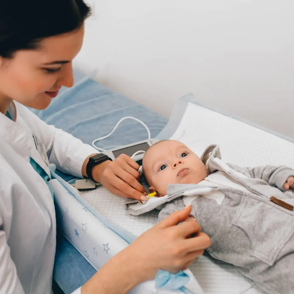 A healthcare professional checks a babys hearing using an ear probe while the baby lies on an exam table, looking up. Medical equipment is in the background.