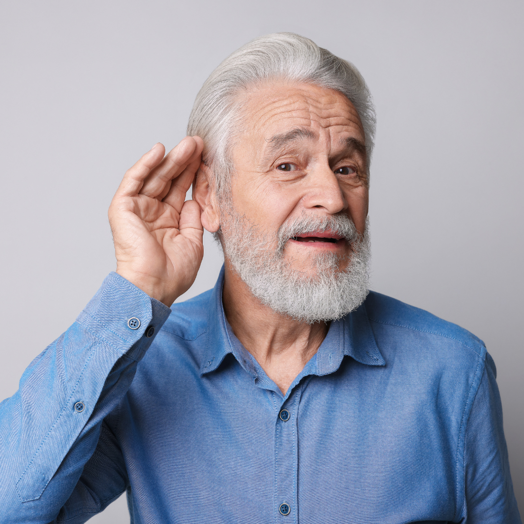 Older man with gray hair and beard wearing a blue shirt, holding his hand to his ear and tilting his head, as if trying to hear something better, against a plain light background.